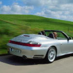996-gen Porsche 911 Buyer Guide: Seen here is a 996-gen Porsche 911 Carrera 4S arctic-silver Cabriolet, from right-rear angle, at speed on a picturesque country road with a pretty woman in the passenger seat. Credit: Porsche AG