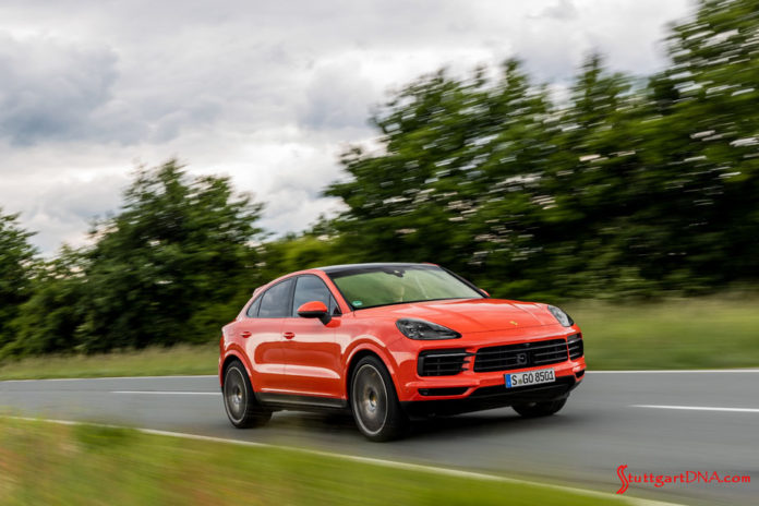 November 2019 Porsche USA sales: This is a photograph of a lava-orange Cayenne coupe, right-front, at speed on a deserted road, with tall green bushes and a cloud-filled sky in b.g. Credit: Porsche AG