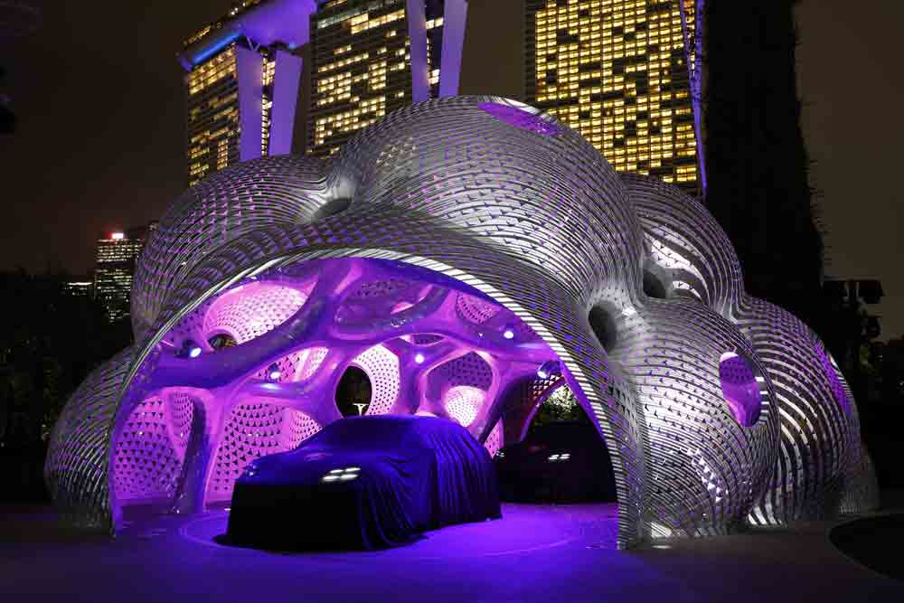 Porsche Macan EV: Displayed here is the colorful cityscape image during the Singapore World Premiere Unveiling, with two Macan EVs indeed veiled with car covers, poised beneath a massive, artistic architectural structure. Credit: Porsche AG